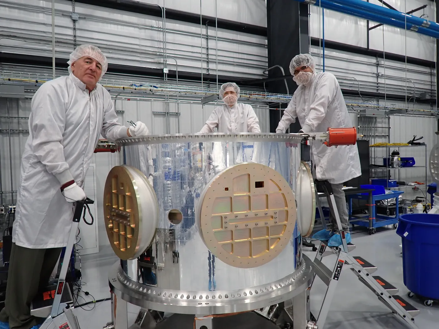 Three technicians in cleanroom garments working around a payload adapter barrel, its circular payload interface plates exposed.