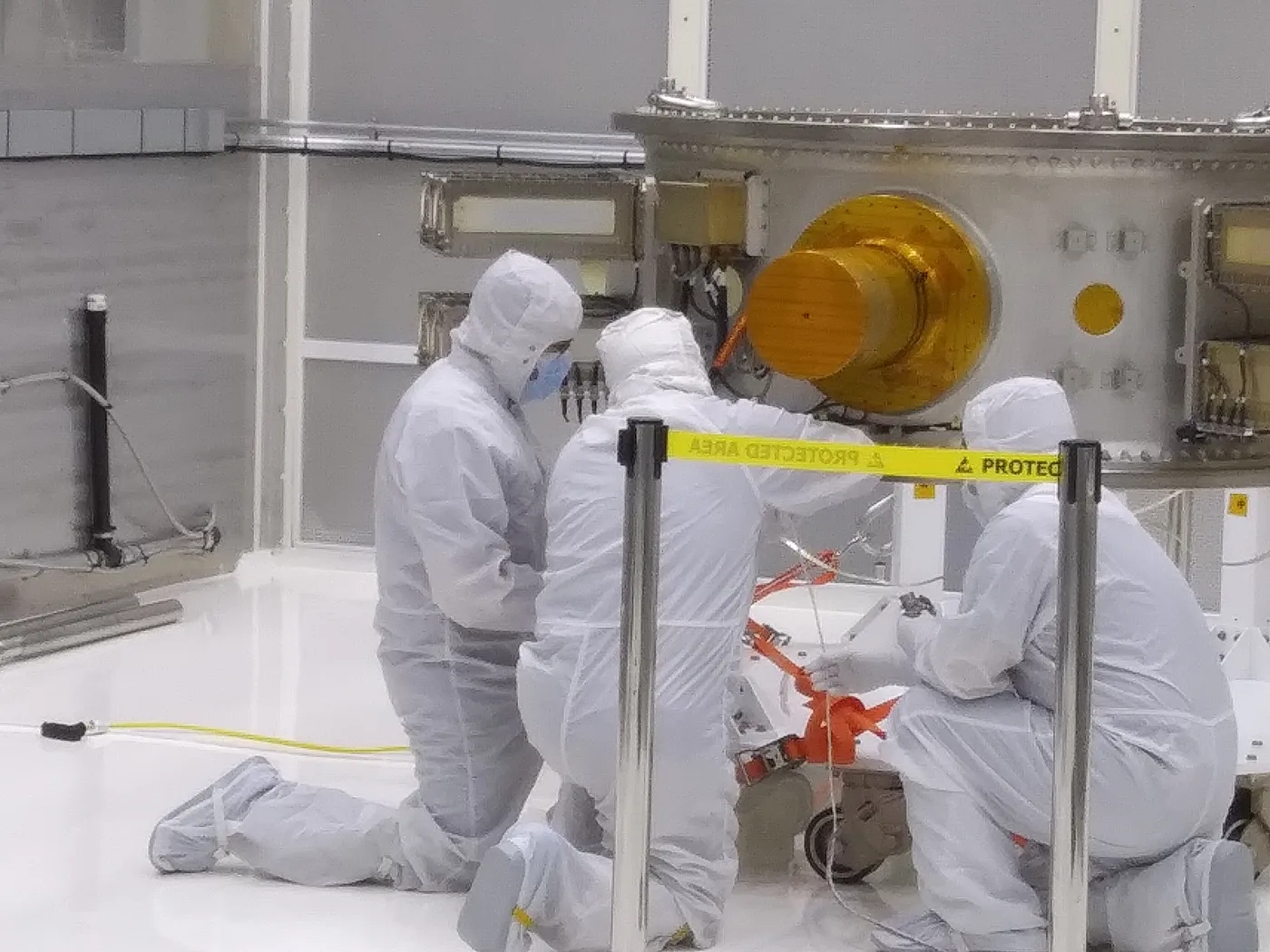Technicians in full cleanroom suits kneeling beside a payload adapter during integration, a gold-covered component mounted on its face.