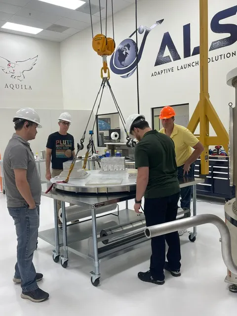 An ALS team gathered around an adapter deck on a handling table beneath a lifting hoist, the ALS logo on the wall behind them.
